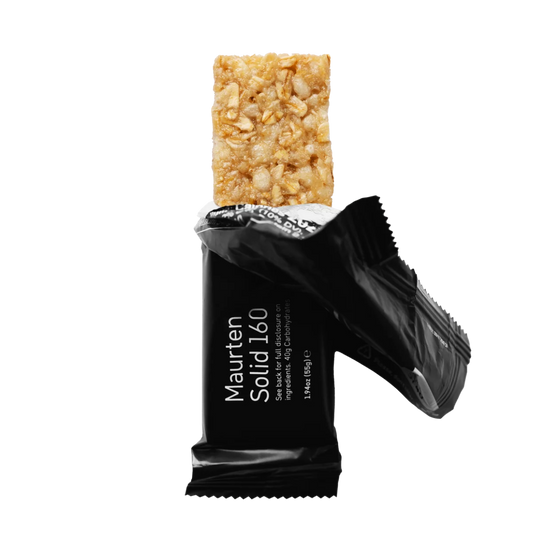 A Maurten Solid 160 energy bar shown with its black wrapper open, revealing a textured, granola-like bar in a warm beige-brown color, set against a solid white background.
