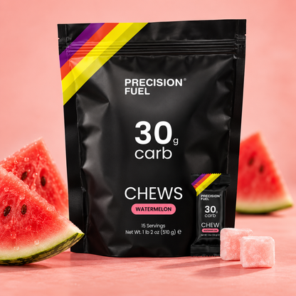 A black stand-up pouch of Precision Fuel 30g carb chews in Watermelon flavor, styled with watermelon slices and pink chewable tablets on a pink background.
